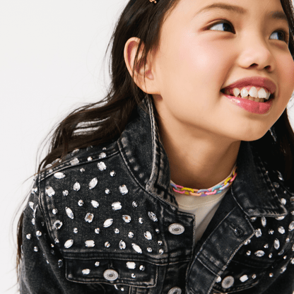 A child smiling and wearing a black denim jacket with white patches and dots.