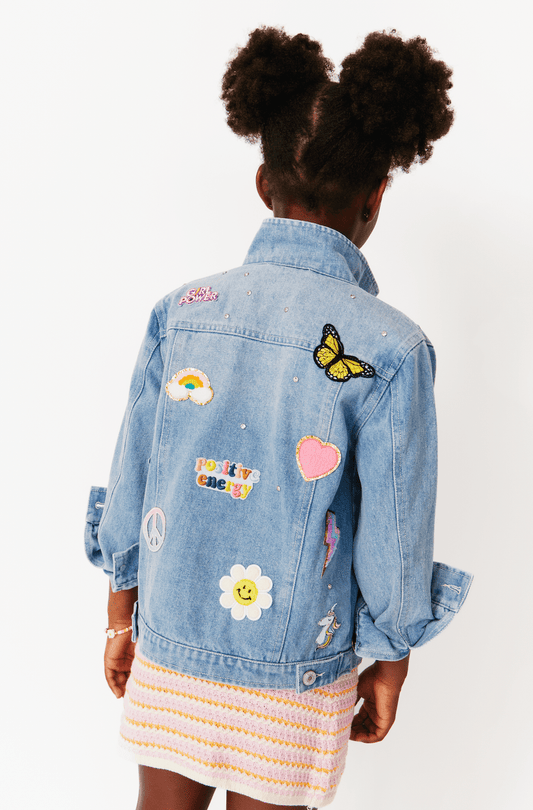 A girl facing away from the camera wears a light wash denim jacket with colorful patches and sequin details.