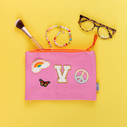 A pink pouch with various patches on it, including a rainbow, a heart, a 'V' for Virginia, and a peace sign. The pouch is laid flat and there are glasses and a beaded bracelet placed on top of it, along with a makeup brush.