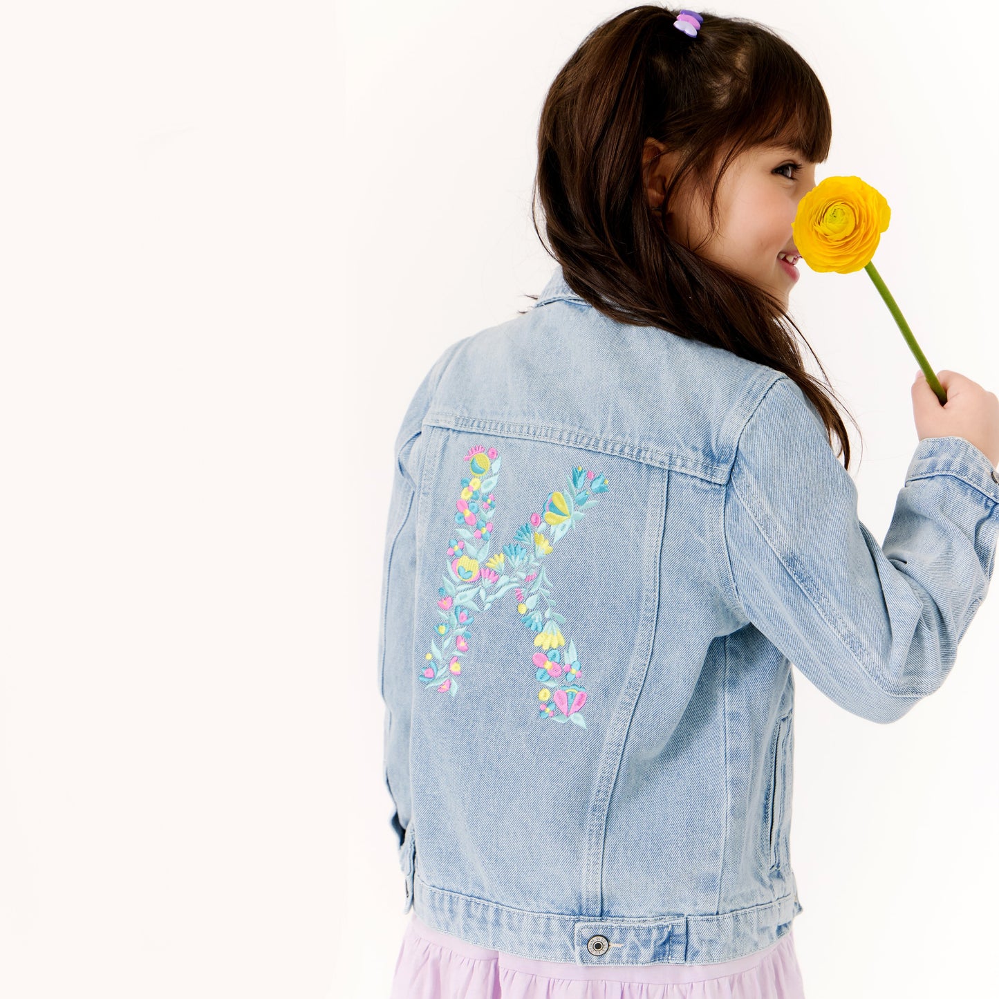 Child wearing a denim jacket with floral embroidery, holding a yellow flower.