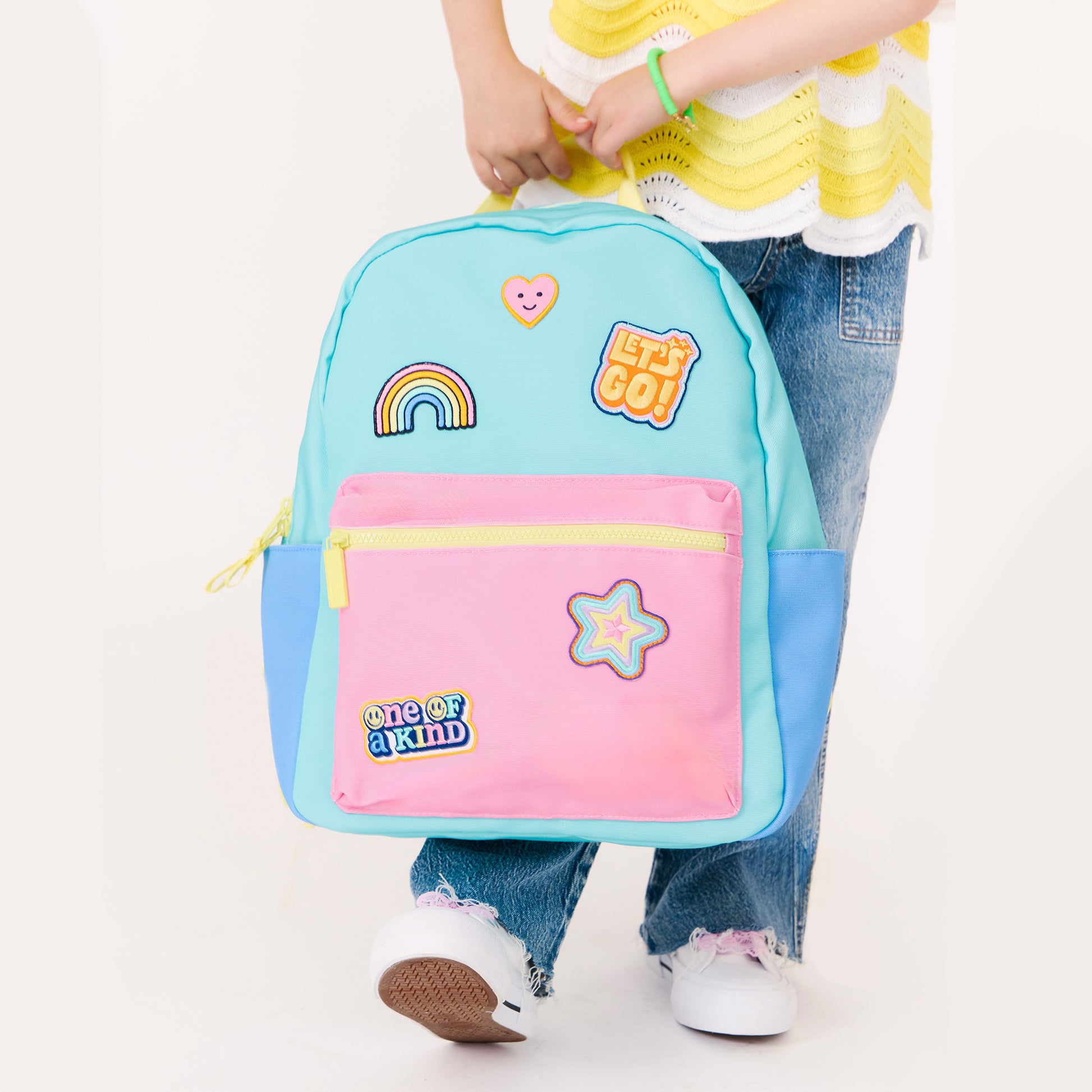 Child holding a pastel blue and pink backpack featuring rainbow, heart, star and colorful patches.