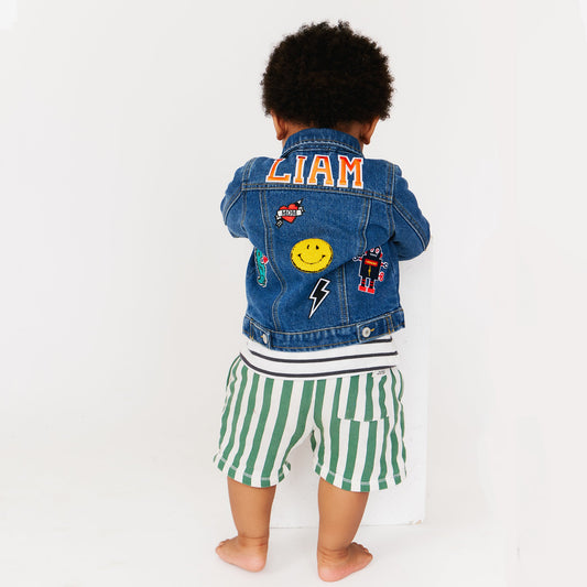 A baby wearing a blue customized denim jacket with various colorful patches on the back. 
