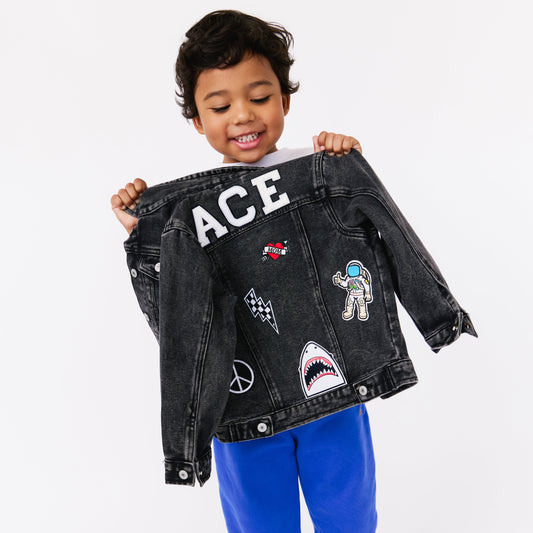 Child holding a black denim jacket with various patches on a white background