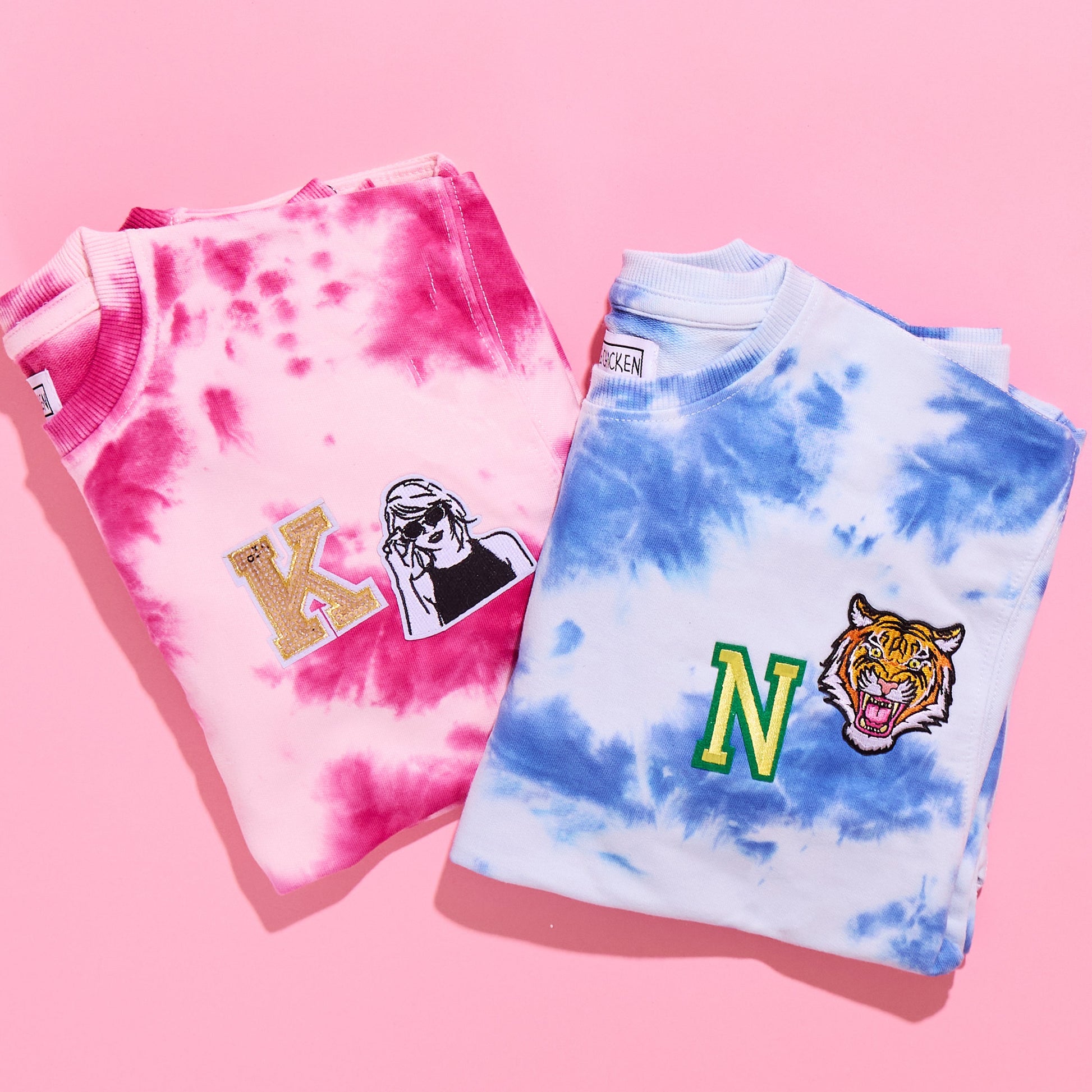 Two tie-dye sweatshirts on a pink background, one is pink with light patterns and the other is blue with darker patterns. Both have personalized patches on them.