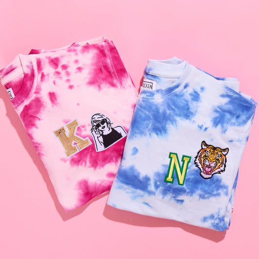 Two tie-dye sweatshirts on a pink background, one is pink with light patterns and the other is blue with darker patterns. Both have personalized patches on them.