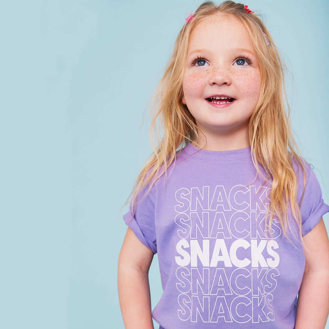 A purple kids' t-shirt with the word 'SNACKS' printed in red letters multiple times.